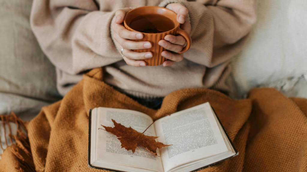 Gemütlich lesen im Herbst: Blatt auf Buch, Kuscheldecke, Hände um Heißgetränk
